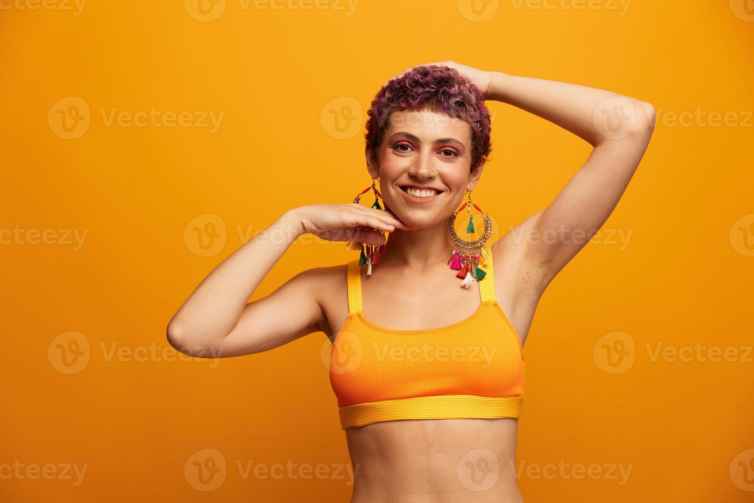 Portrait of a young woman with a short haircut and colored hair smiling and showing her tongue at the camera on an orange background with earrings accessories in the studio photo