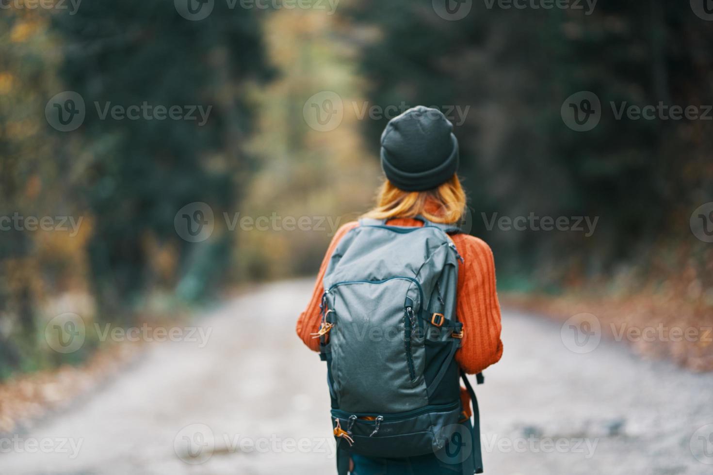 woman with a backpack in a hat and an orange sweater on the road in the autumn forest back view photo