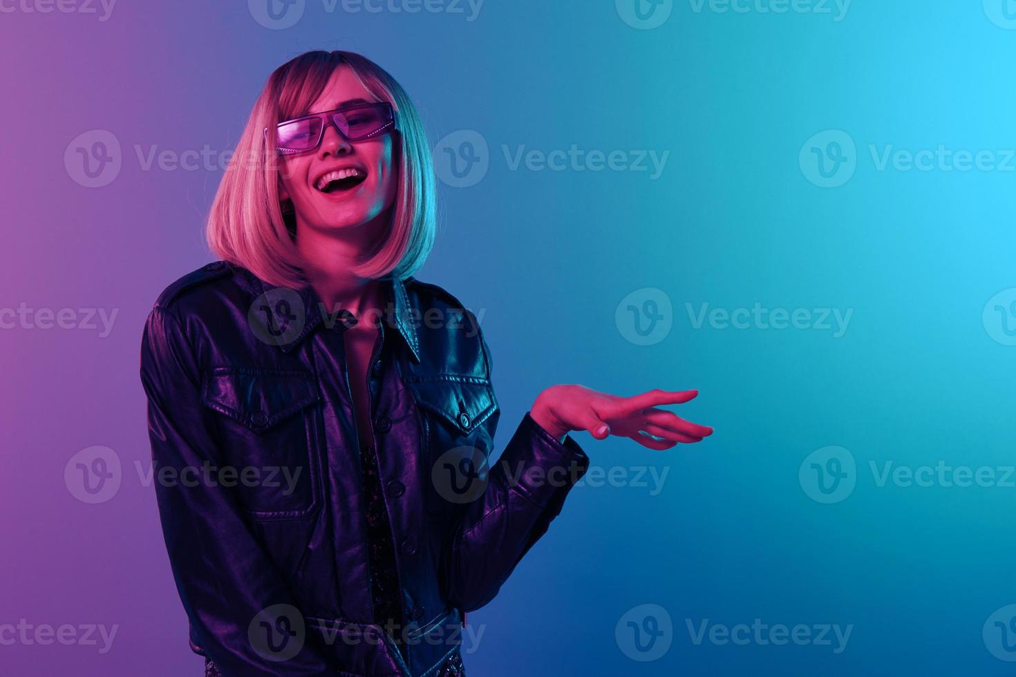 Laughing sexy beautiful blonde woman in leather jacket sparkly dress trendy sunglasses look aside posing isolated in blue pink color light studio background. Neon party Cyberpunk concept. Copy space photo