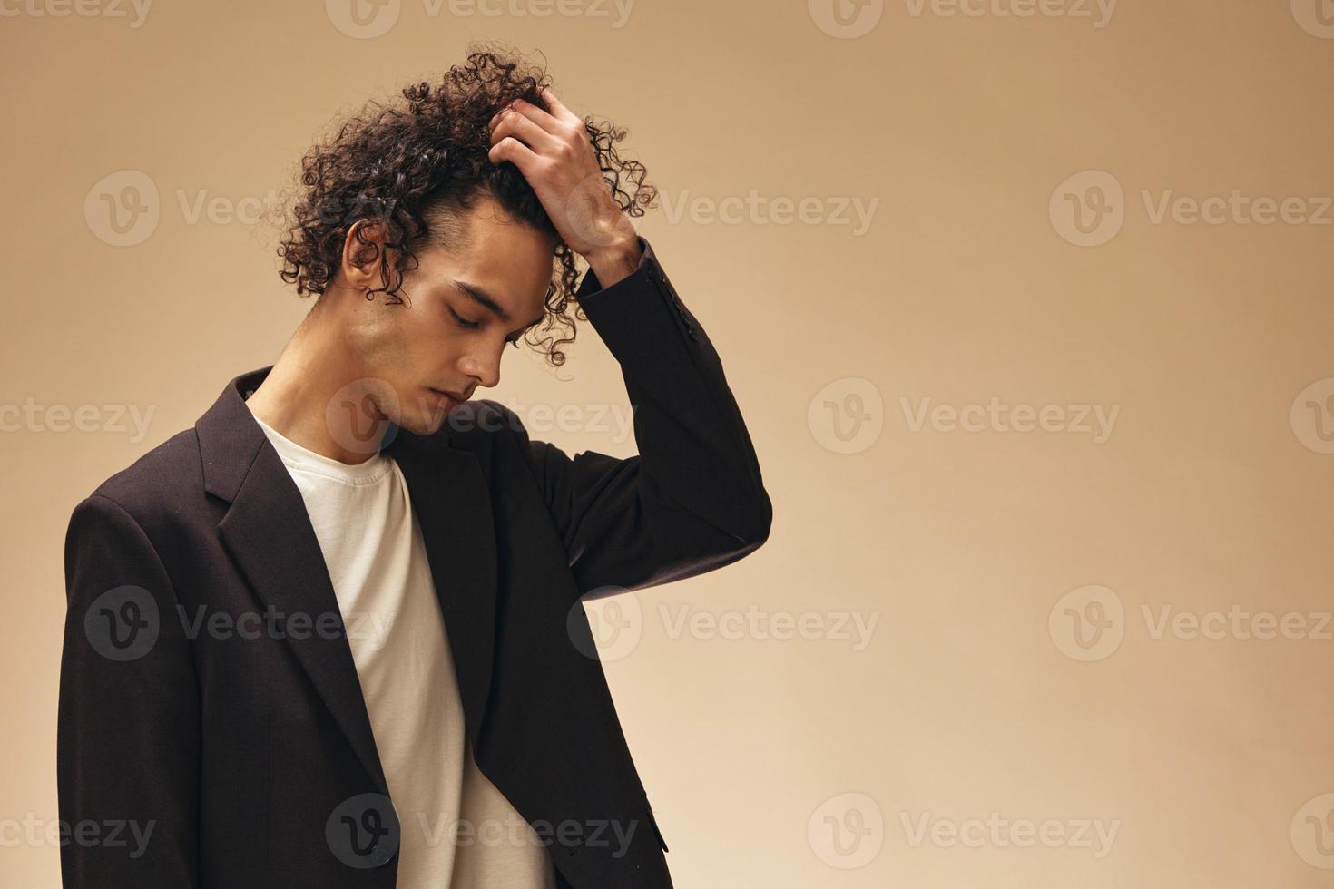 Defiant awesome tanned curly man in classic brown jacket touches curls posing isolated on over beige pastel background. Fashion New Collection offer. Retro style concept. Free place for ad photo