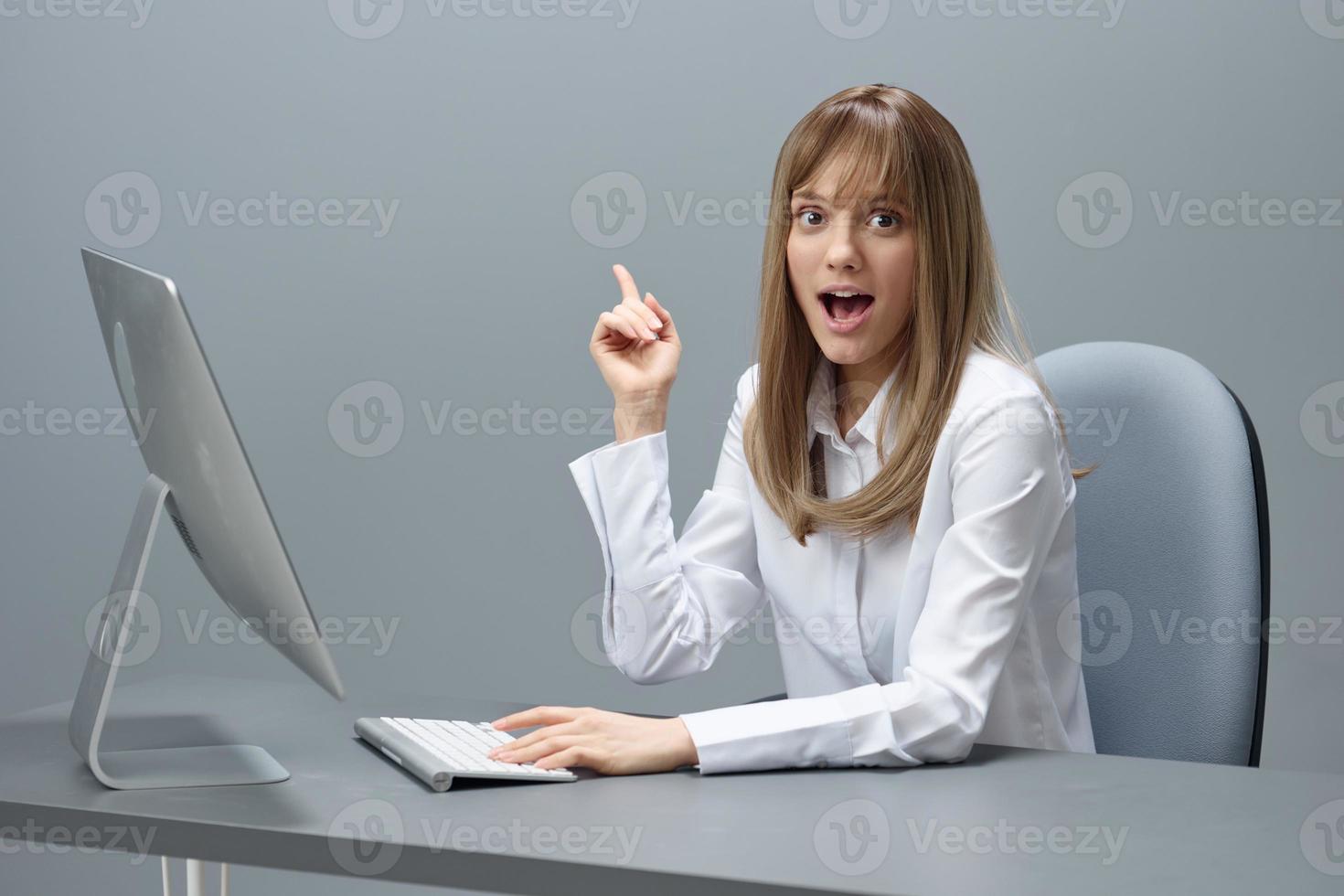 Excited happy blonde businesswoman freelancer have cool idea point finger up using desktop computer sitting at workplace in gray modern office. Remote Job And Career Profession Concept. Copy space photo