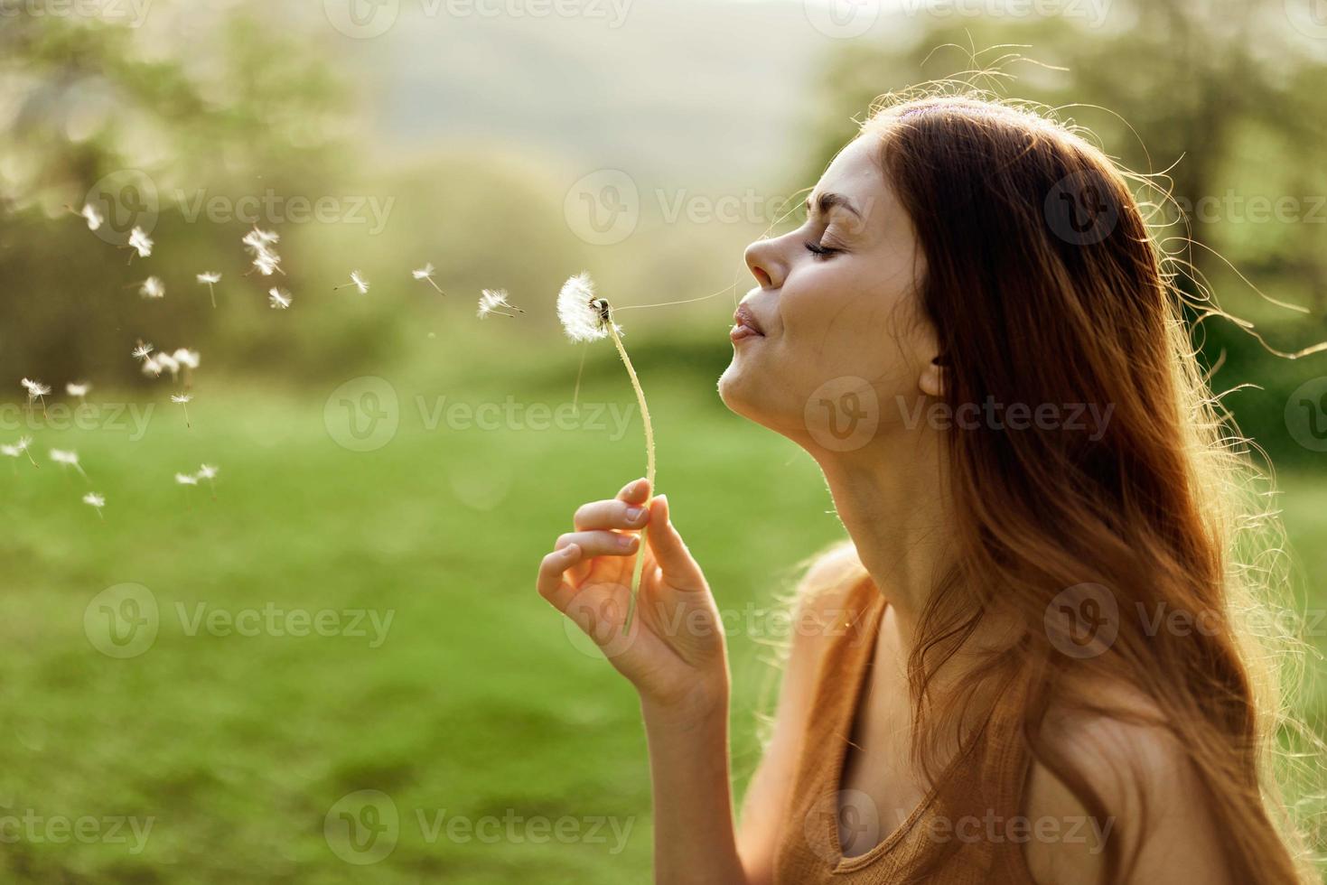 The woman smiles and blows the dandelion seeds into the wind. Summer and sunset light photo