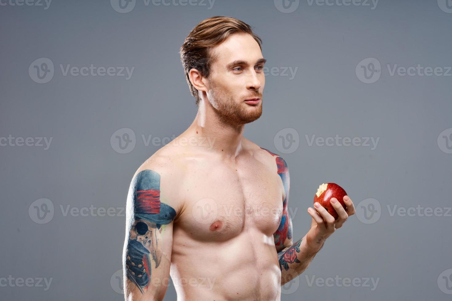 sexy athlete with pumped up muscles eats an apple on a gray background and a tattoo on his arm photo