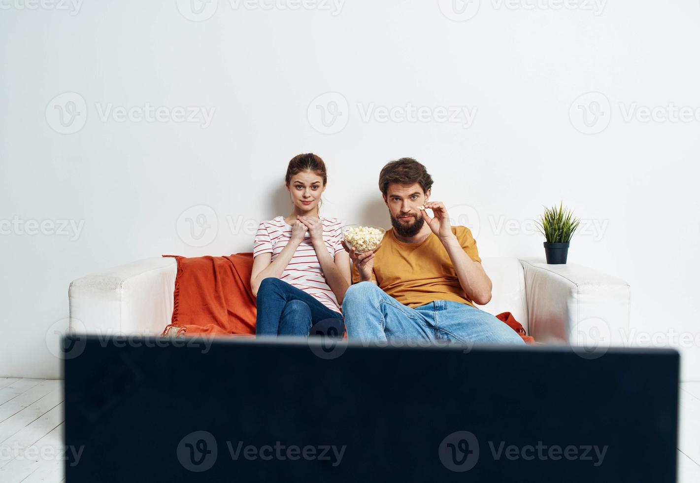 un hombre y un mujer son sentado en el sofá en frente de el televisión y un verde flor en un maceta adentro foto