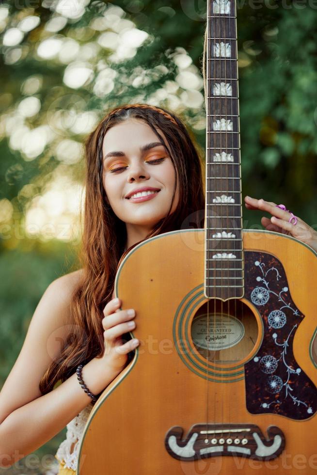 un joven hippie mujer con un guitarra en su manos sonrisas dulcemente dentro el cámara en un viaje a naturaleza estilo de vida en armonía foto