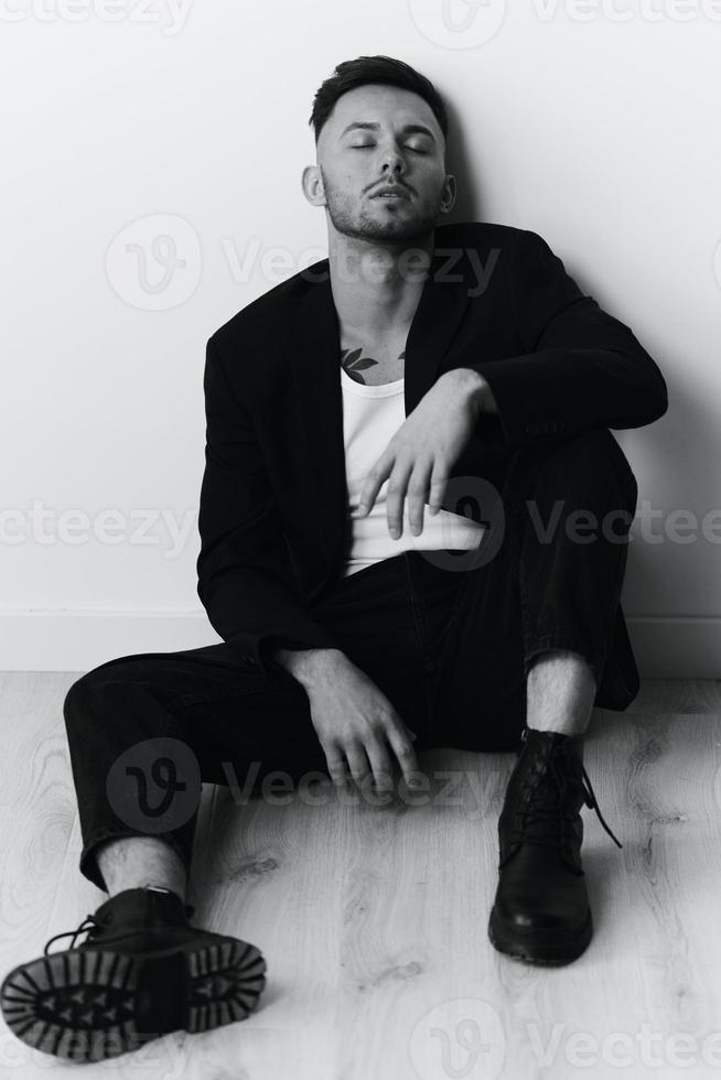 modelado instantáneas grave seguro de sí mismo atractivo hermoso hombre en negro chaqueta sentado en piso posando en blanco estudio antecedentes. negro y blanco concepto. Copiar espacio foto