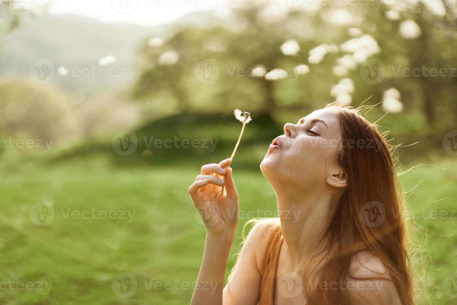 el contento mujer sonrisas y golpes el diente de león en el viento. verano verde paisaje y Brillo Solar en el antecedentes foto