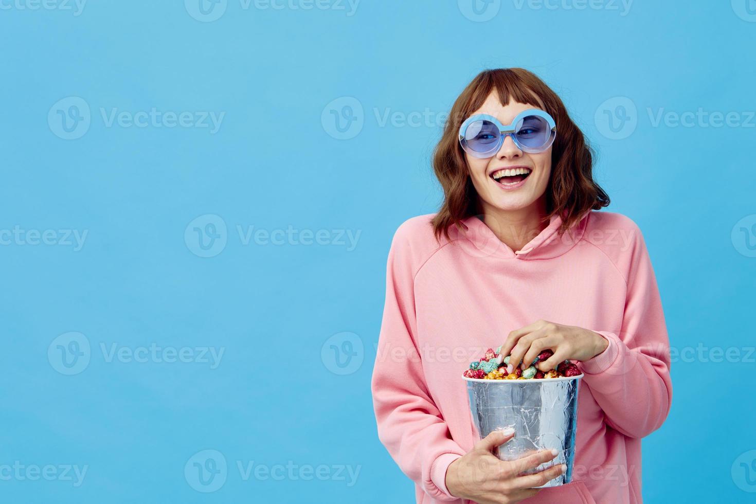 Evening movie concept. Lovely smiling cute redhead lady in pink hoodie sunglasses with popcorn posing isolated on blue studio background. Copy space Banner. Fashion Cinema. Entertainment offer photo