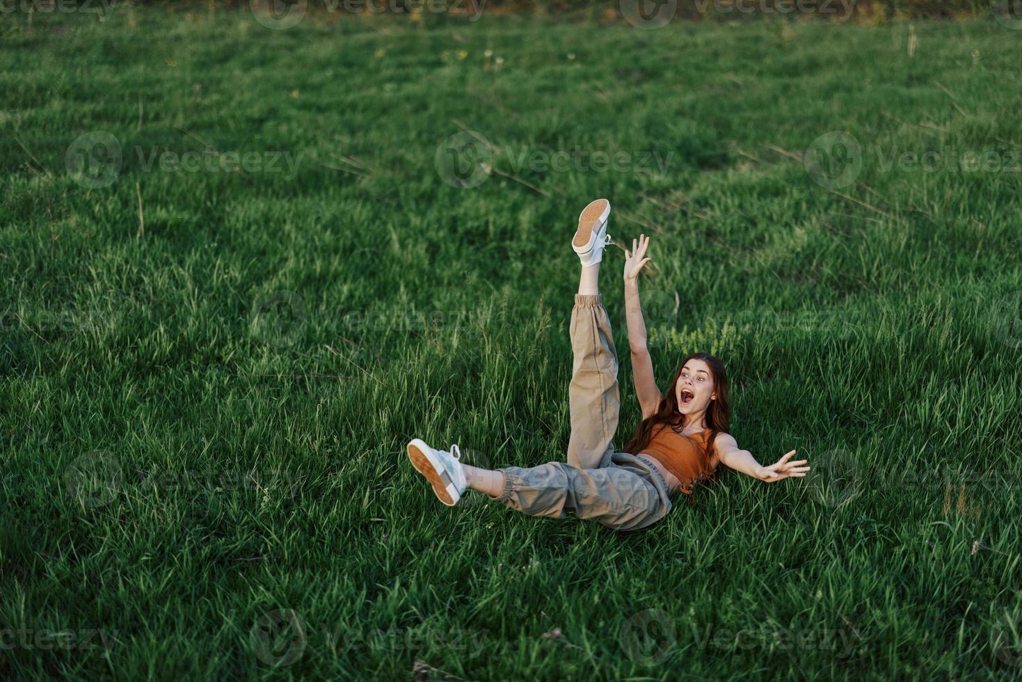 A young woman playing games in the park on the green grass spreading her arms and legs in different directions falling and smiling in the summer sunlight photo