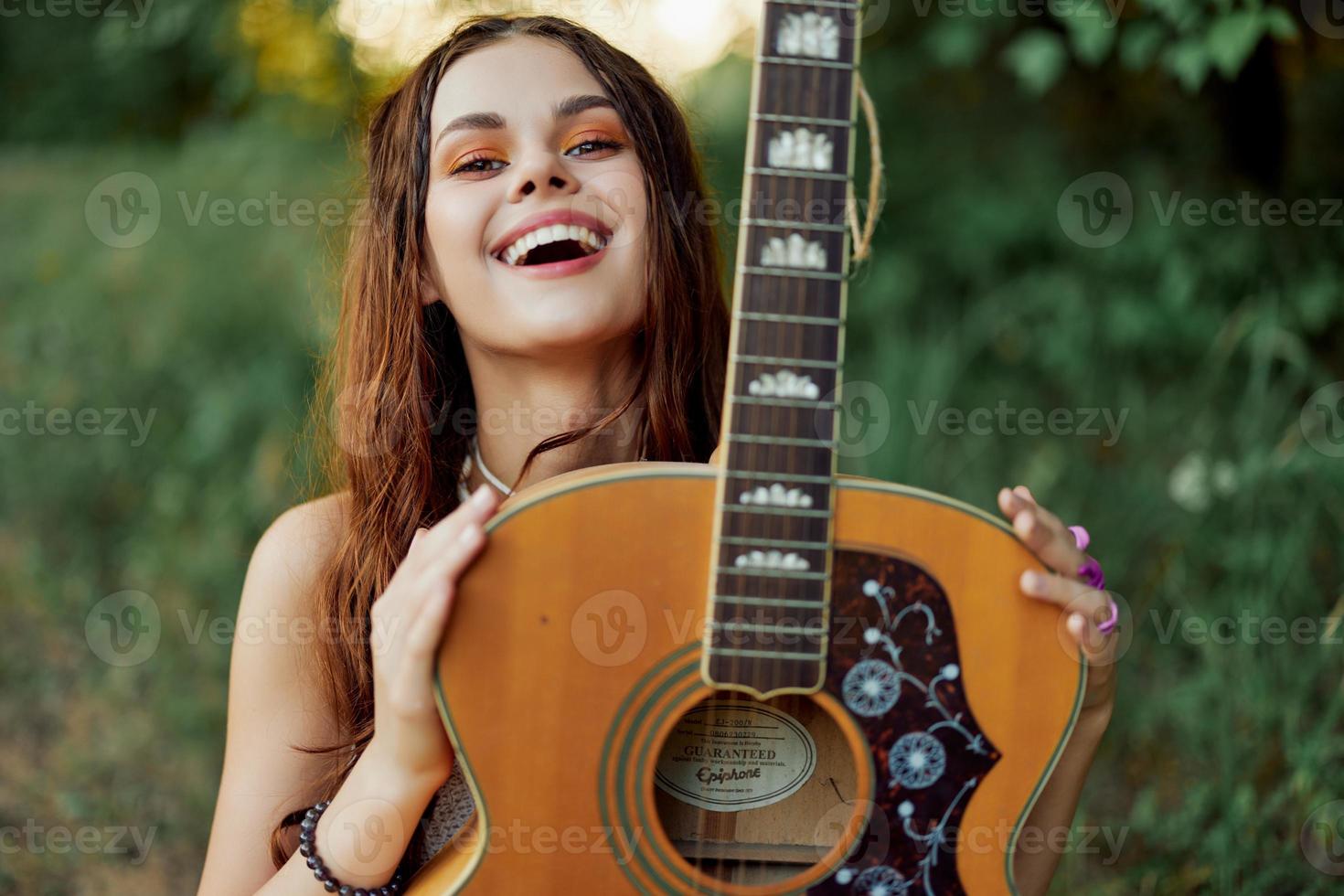 joven hippie mujer con eco imagen sonriente y mirando dentro el cámara con guitarra en mano en naturaleza en un viaje foto