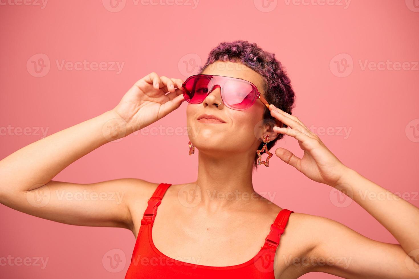 Fashion portrait of a woman with a short haircut in colored sunglasses with unusual accessories with earrings smiling on a pink bright background with a fitness body dancing photo