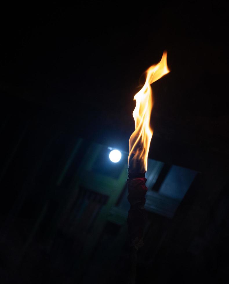 A burning torch at night time with yellow flames. Ancient wooden torch burning on a dark background. A torch burning in the night and a white light glowing in the background. Torch with yellow flame. photo