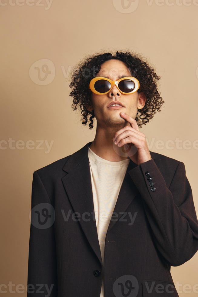 Demonstrative awesome tanned curly man in classic brown jacket trendy sunglasses posing isolated on over beige pastel background. Fashion New Collection offer. Retro style concept. Free place for adDemonstrative awesome tanned curly man in classic brown j photo