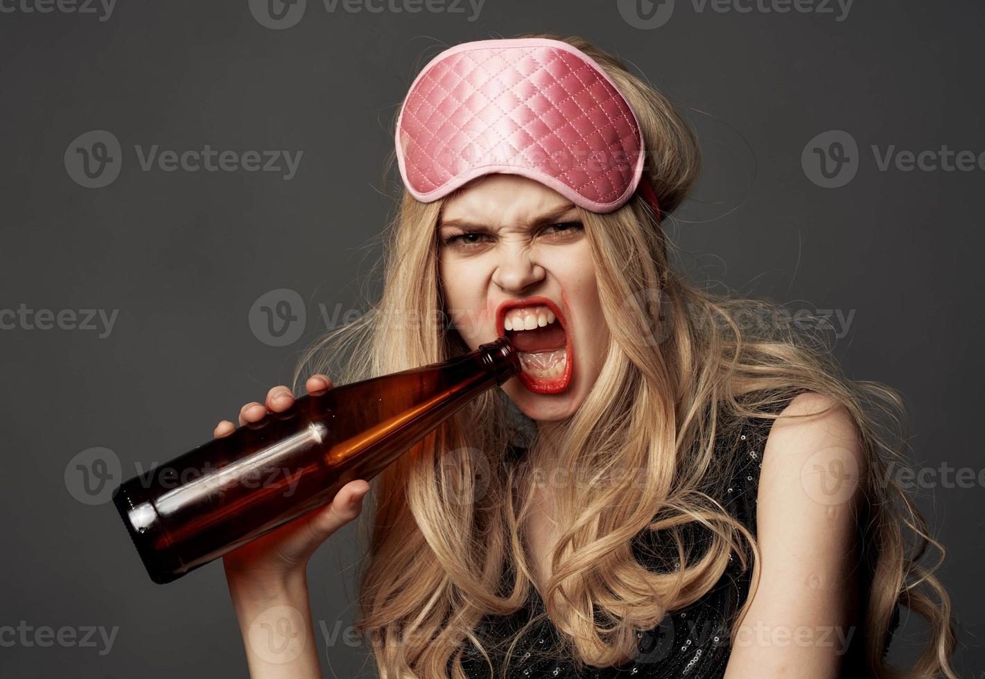 enojado mujer con un botella de cerveza en su mano con brillante maquillaje y un rosado dormir máscara foto