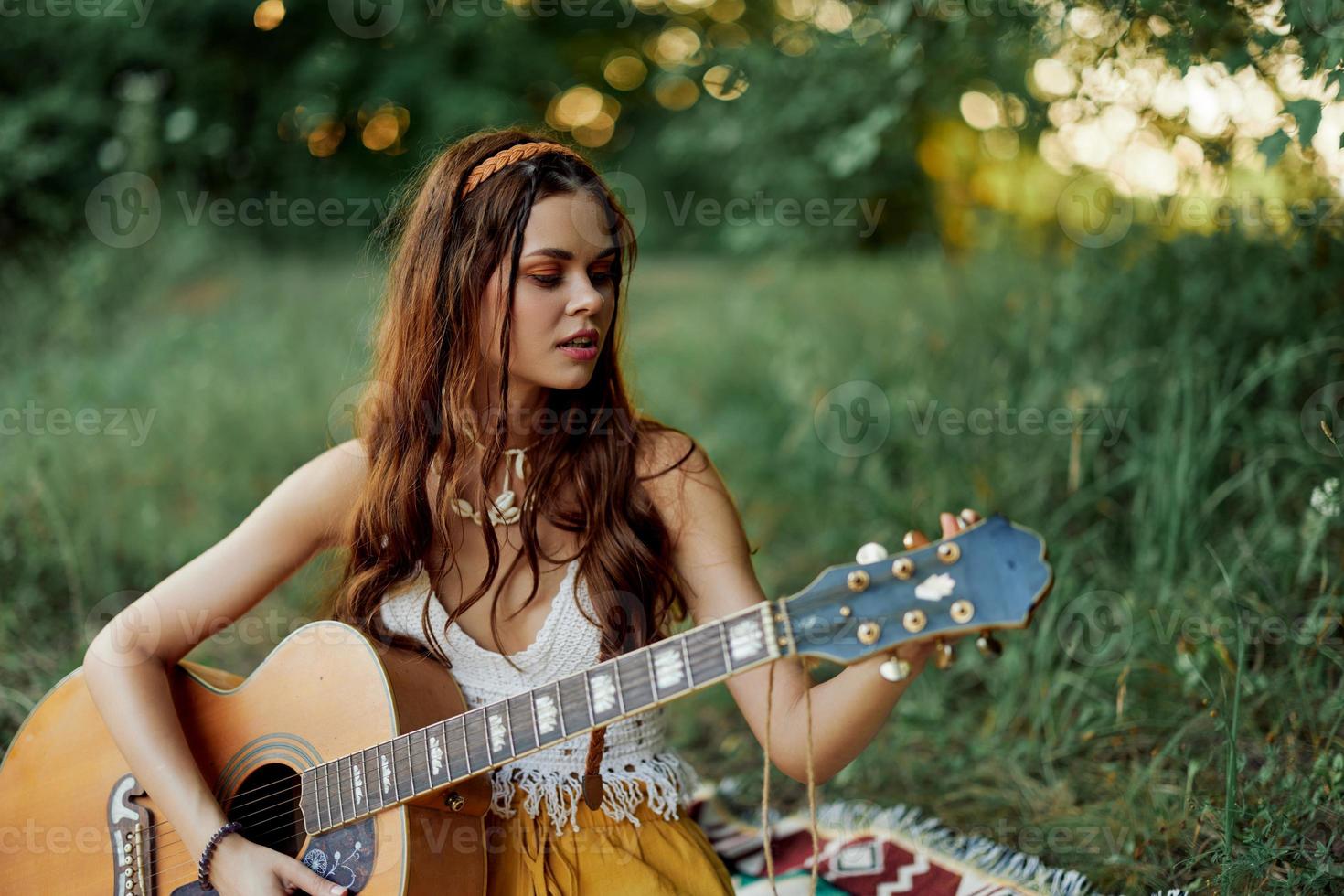 Girl hippie woman playing guitar in eco-friendly clothes sitting on the ground outside in nature in the fall watching the sunset photo