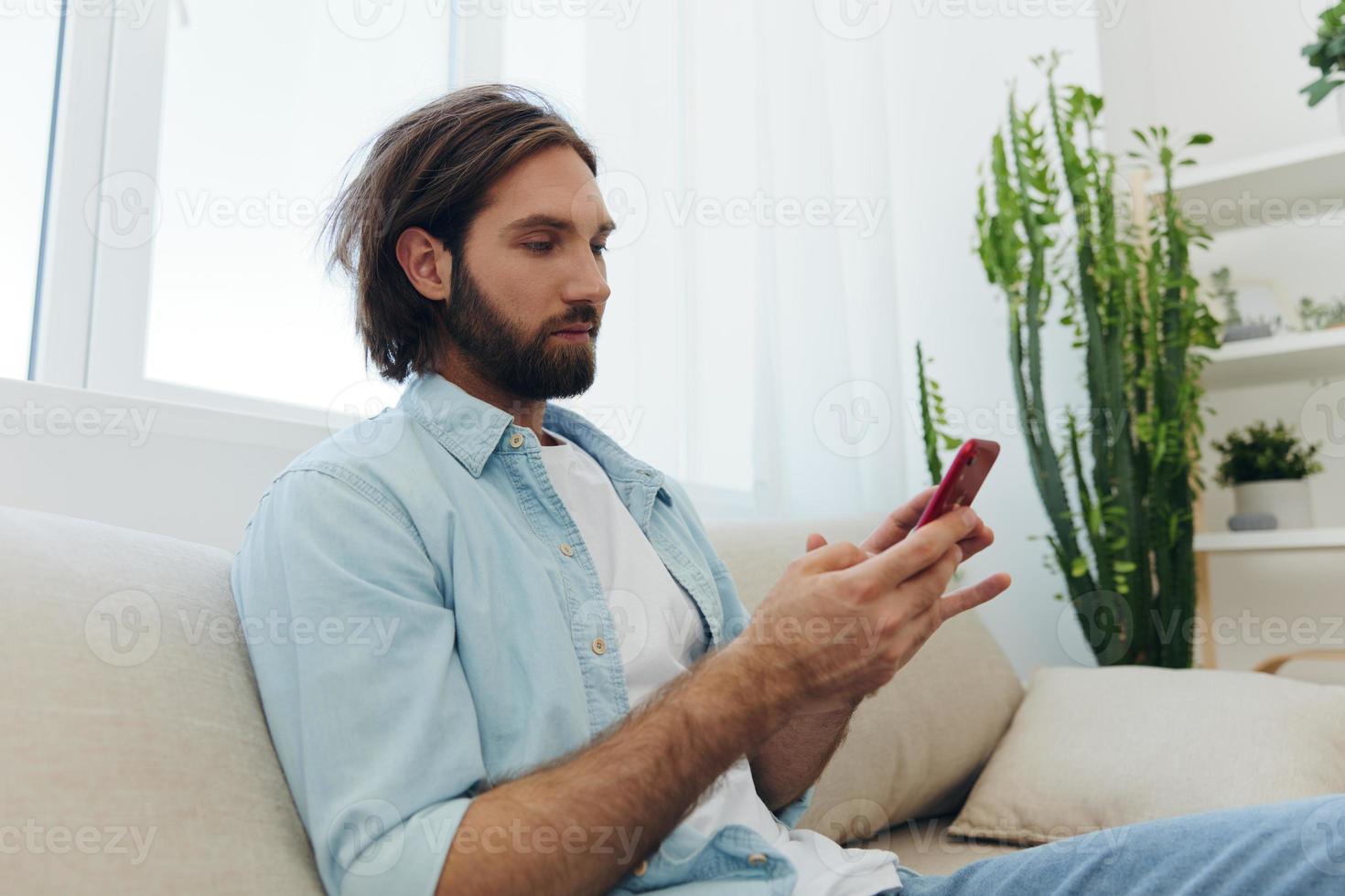 A freelance man in a white T-shirt, blue jeans, and shirt sits on the couch with his phone at home on his day off and plays games photo