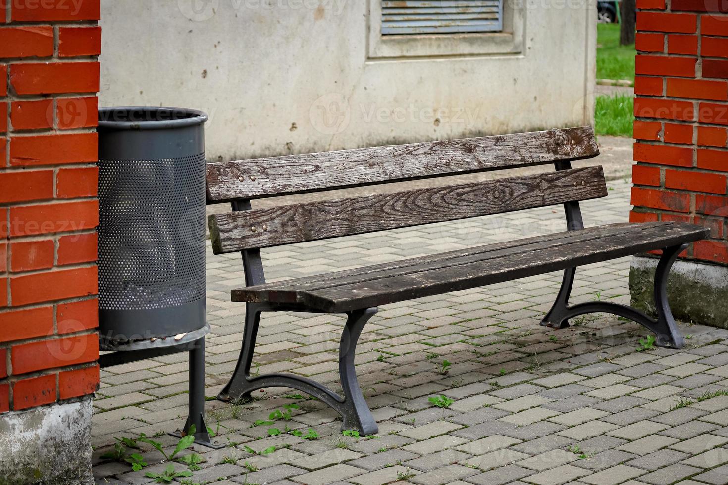 Brown wooden bench with black metal legs standing in a yard between two building columns of red brick near a black trash bin photo