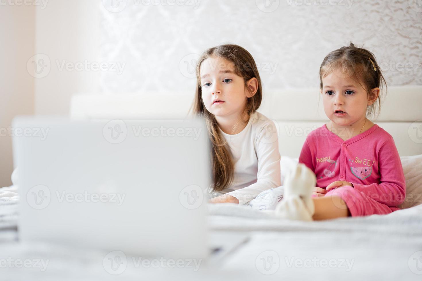 Two cute little sisters wear pajamas looking cartoon by laptop in morning at home. photo