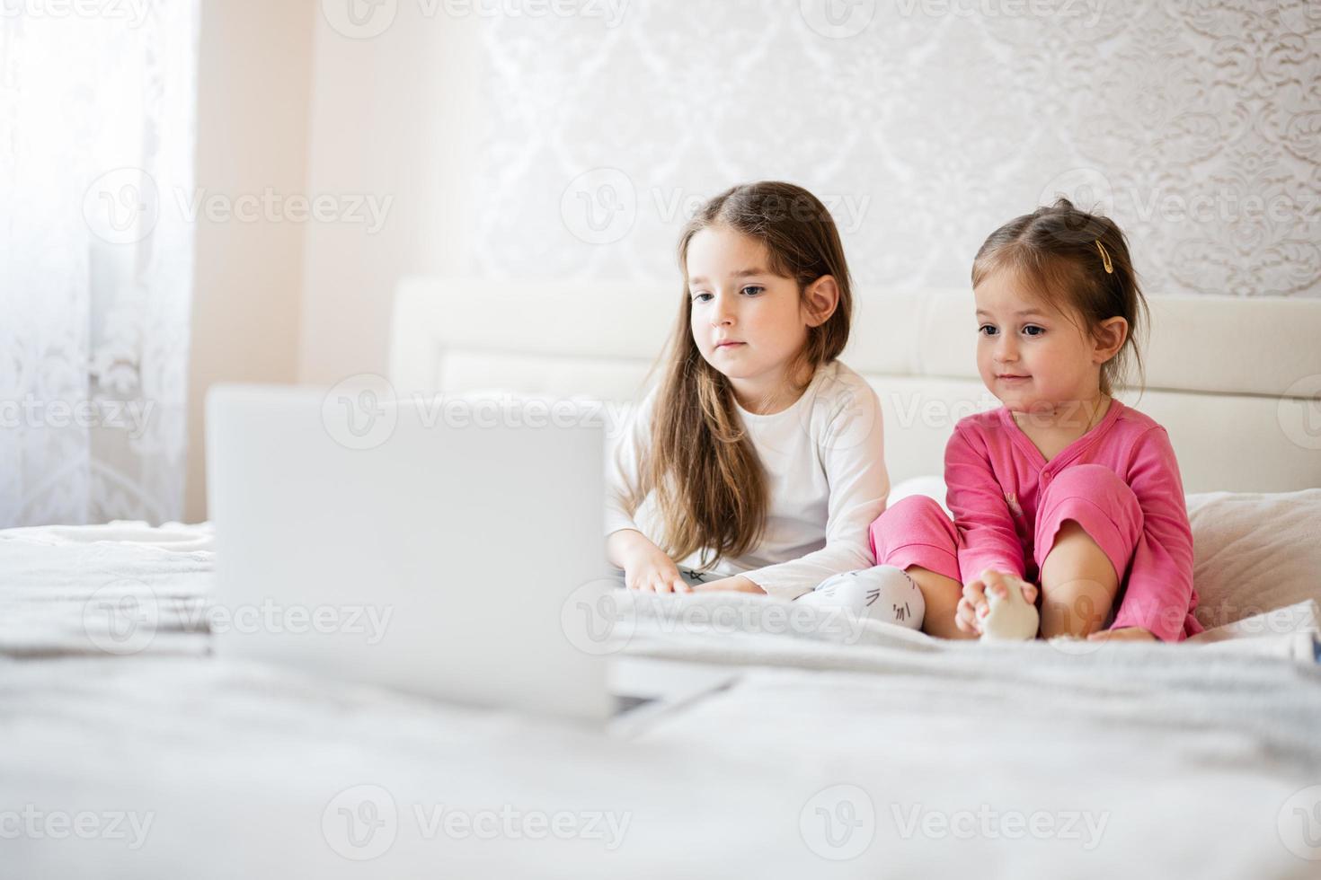 Two cute little sisters wear pajamas looking cartoon by laptop in morning at home. photo