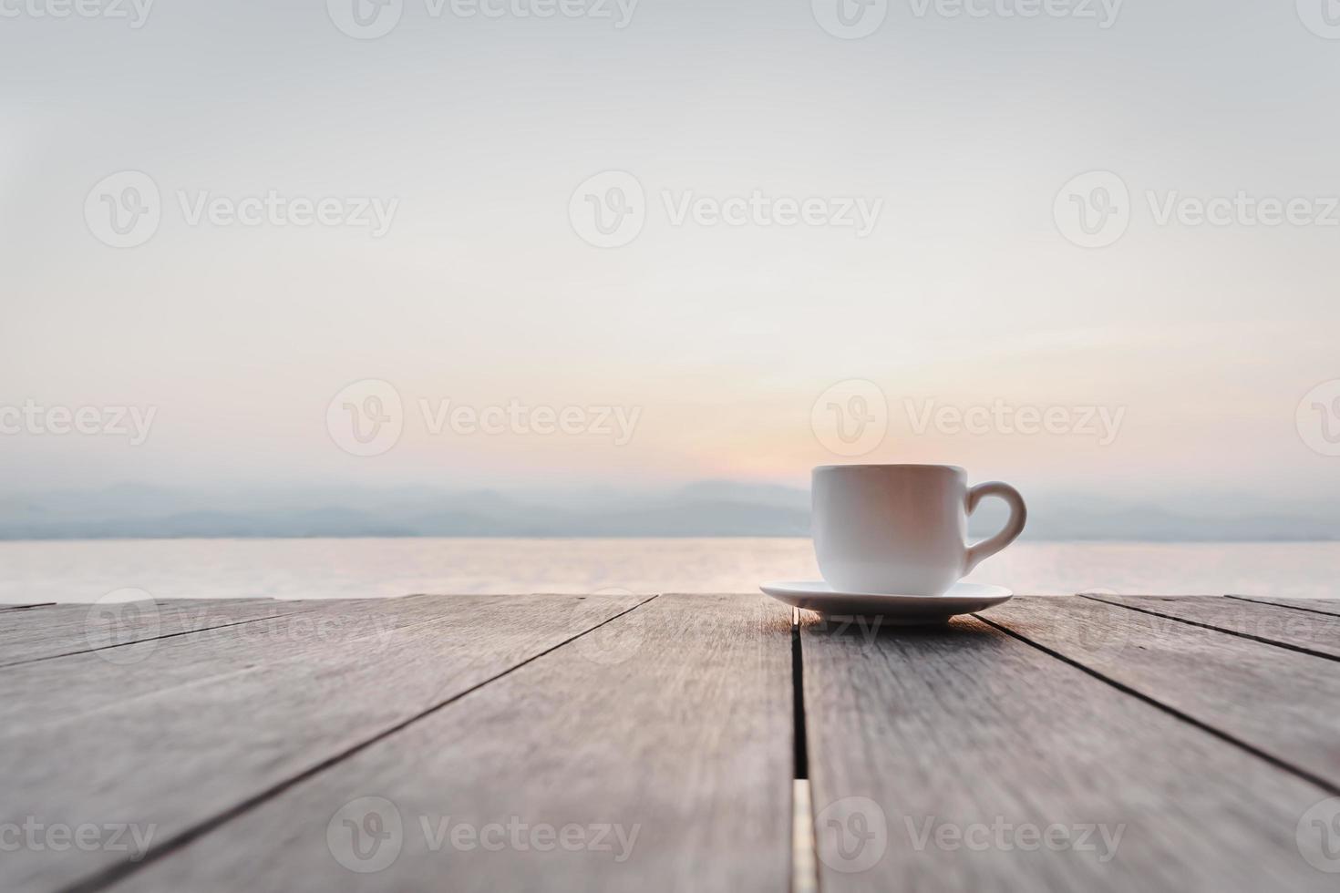 Cup of coffee on wooden deck over sunrise with river mountain in background. photo