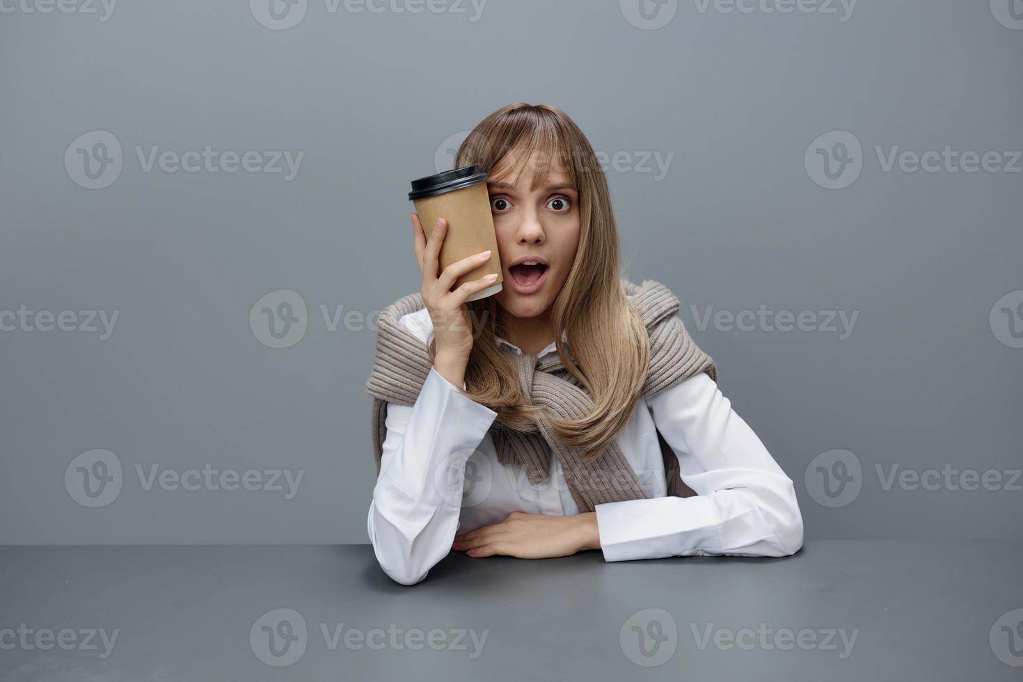 Shocked happy young blonde student lady freelancer in warm sweater with takeaway coffee sitting open mouth in gray modern home office. Coffee break Lover Concept. Copy space. Cool offer photo