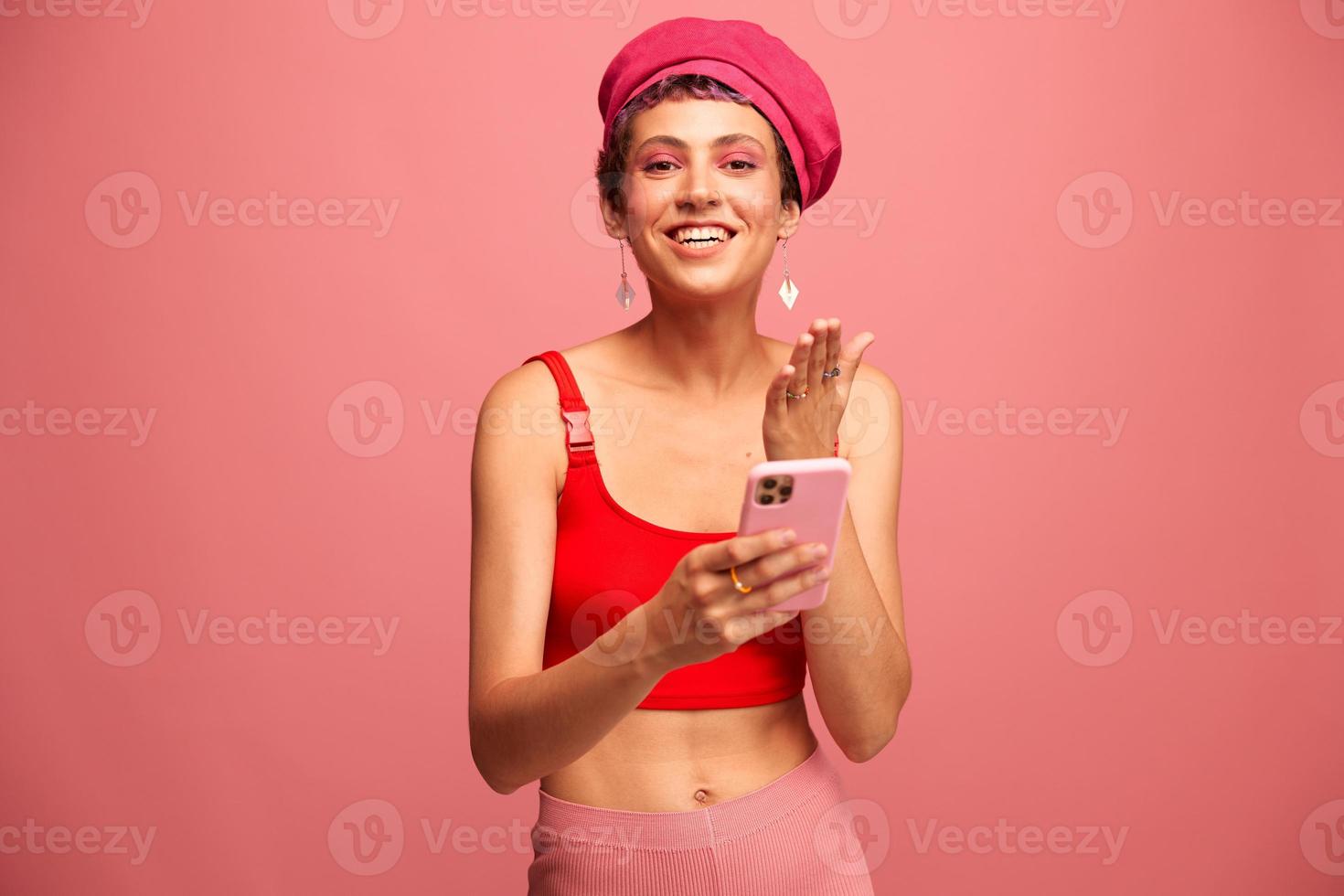 Young woman blogger with colored pink hair and short haircut flipping through the phone screen and typing a message with a smile and surprise in stylish clothes on a pink background photo