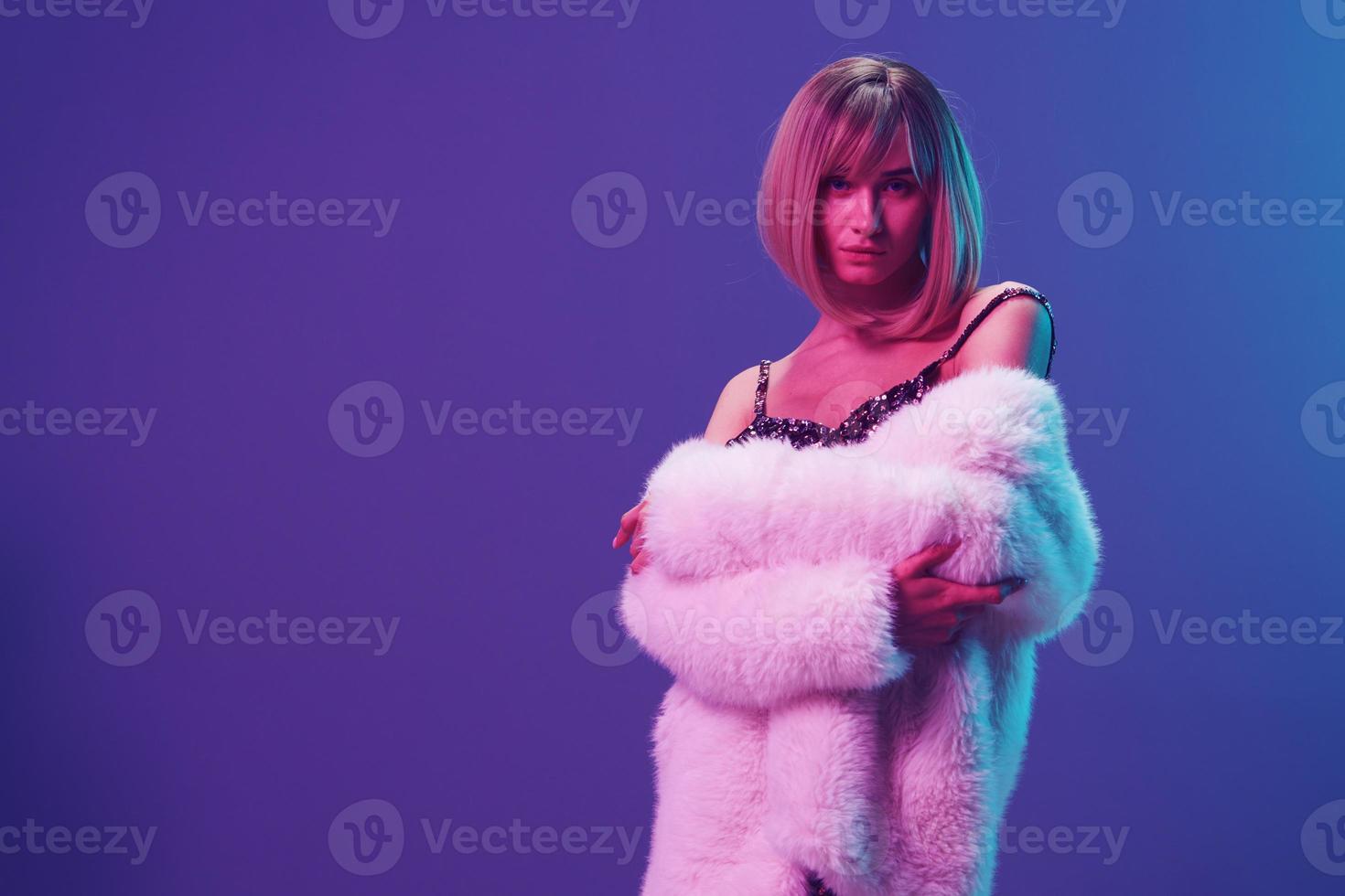 Sexy blonde woman in fluffy fur coat sparkly dress with open shoulders cross hands posing isolated in blue violet pink color light studio background. Neon party Fashion concept. Copy space Banner photo