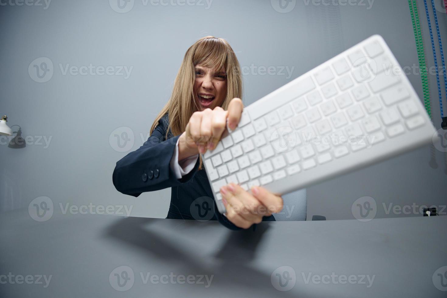 loco irritado enojado milenario rubia mujer de negocios trabajador en azul chaqueta lanza teclado y gritando a cámara en gris moderno oficina. personas emociones negocio concepto. Copiar espacio, amplio ángulo foto