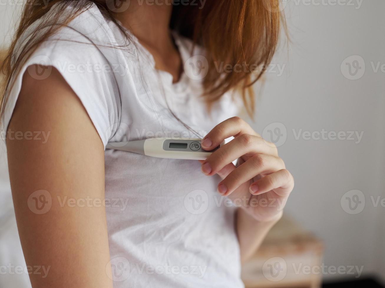 un mujer en un blanco camiseta es comprobación el temperatura con un termómetro debajo su brazo foto