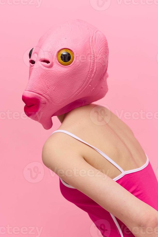 Skinny woman in a pink Halloween costume with a fish head on her face poses funny against a pink background and looks at the camera, art concept photo