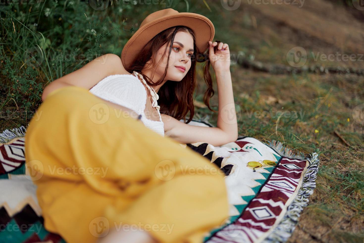 A woman in eco-friendly clothing in a hippie look is lying on a colored plaid smiling and looking at an autumn sunset in nature in the park. Lifestyle on a journey of harmony with the world photo