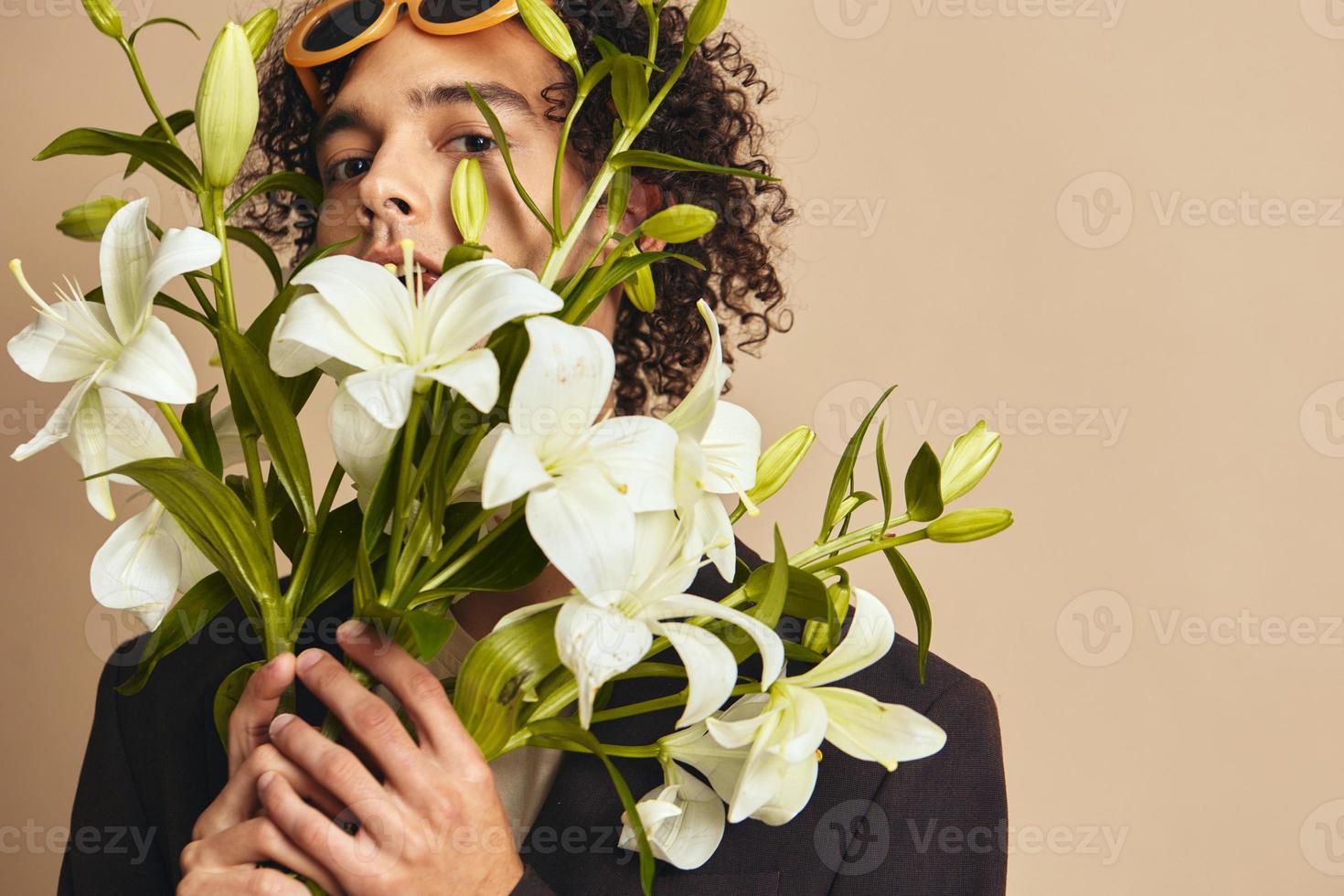 Closeup portrait of handsome tanned curly man in trendy sunglasses hold lilies near face posing isolated on over beige background. Fashion New Collection offer. Retro style concept. Free place for ad photo