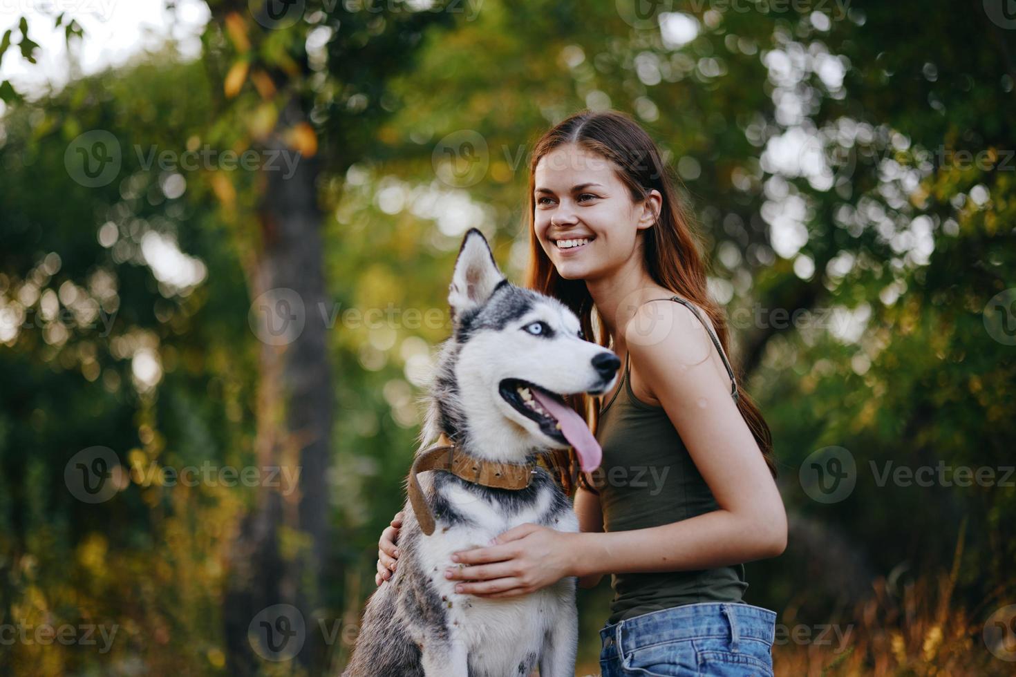 A woman with a husky breed dog smiles and affectionately strokes her beloved dog while walking in nature in the park in autumn against the backdrop of sunset photo