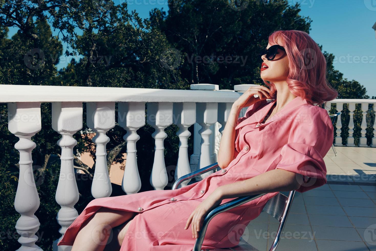 a glamorous woman in sunglasses in the summer sits on the street on a chair near the railing photo