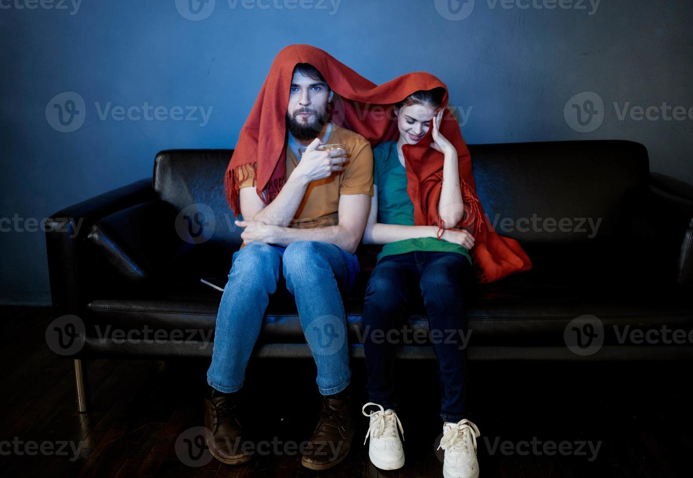 a woman and a man under a red blanket on the couch watching tv in the evening photo