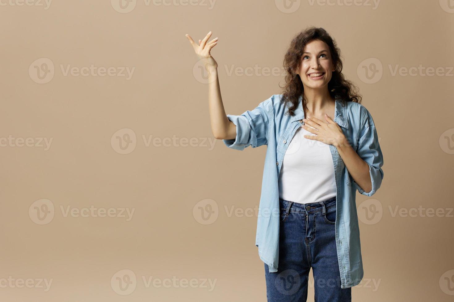 Excited happy enjoyed curly beautiful female in jeans casual shirt point hands looks up at free place posing isolated on over beige pastel background. People Lifestyle emotions concept. Copy space photo