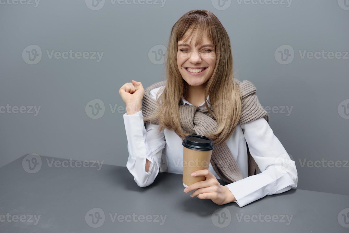 Overjoyed cute young blonde student lady freelancer in warm sweater with takeaway coffee raise fist up smiling close eyes in gray modern home office. Coffee break Lover Concept. Copy space. Cool offer photo