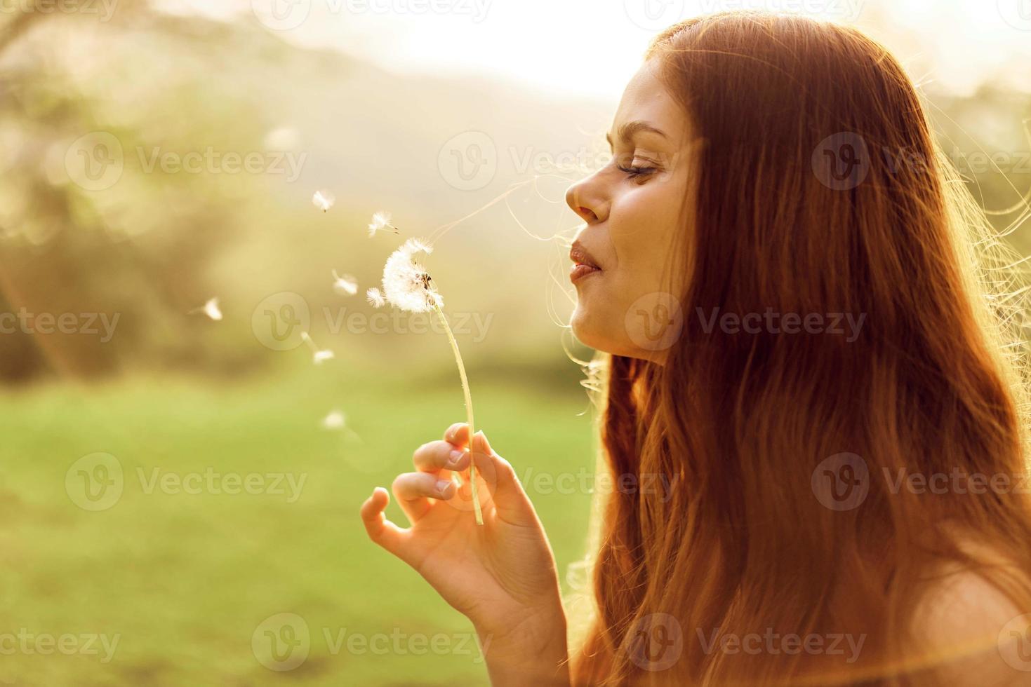Portrait of a young woman in profile with a dandelion flower in her hand blowing on it and smiling against the green summer grass in the setting sun in nature photo