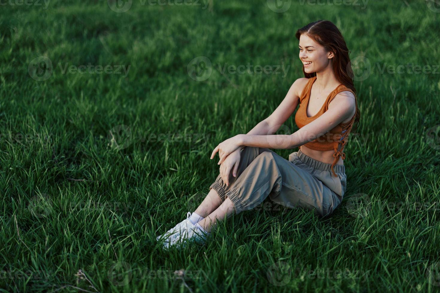 un contento mujer mira fuera a el ajuste verano Dom sentado en el Fresco verde césped en el parque y sonriente, ver desde arriba. el concepto de cuidados personales foto