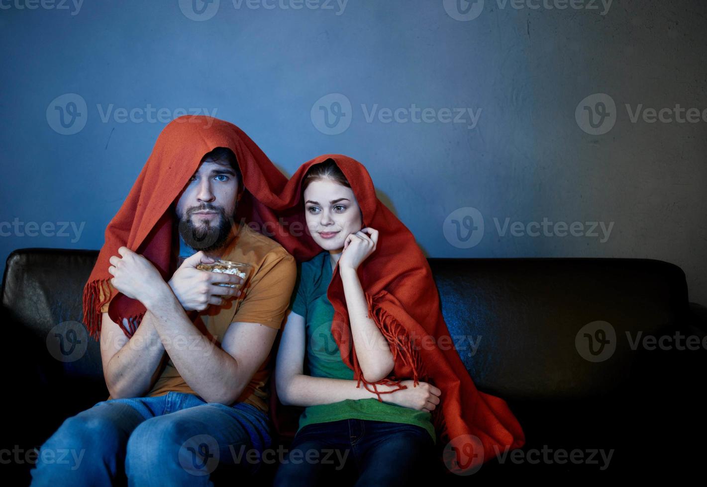 a woman and a man under a red blanket on the couch watching tv in the evening photo