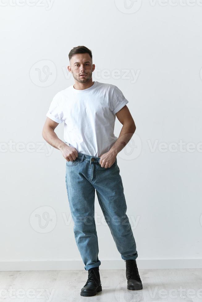 Full body shot of handsome serious tanned man guy in basic t-shirt holds hand on jeans posing on white background. Fashion Style New Collection Offer. Copy space for ad. Modeling snapshots photo