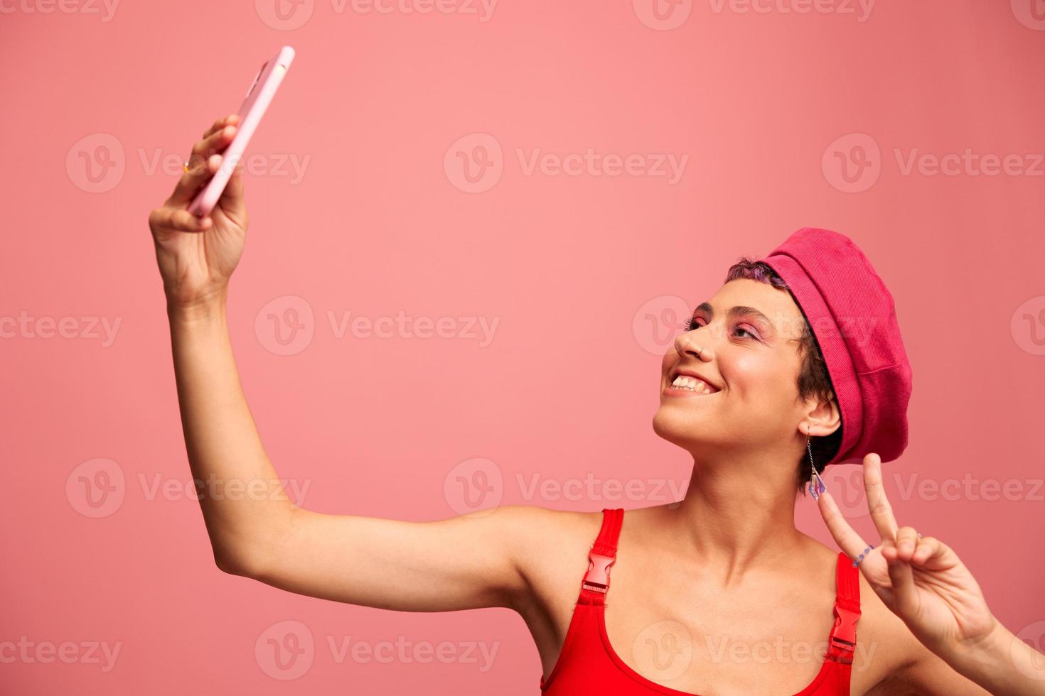 un joven mujer blogger con de colores rosado pelo y un corto Corte de pelo toma un imagen de sí misma en el teléfono y retransmisiones un sonrisa en elegante ropa y un sombrero en un rosado antecedentes monocromo estilo foto