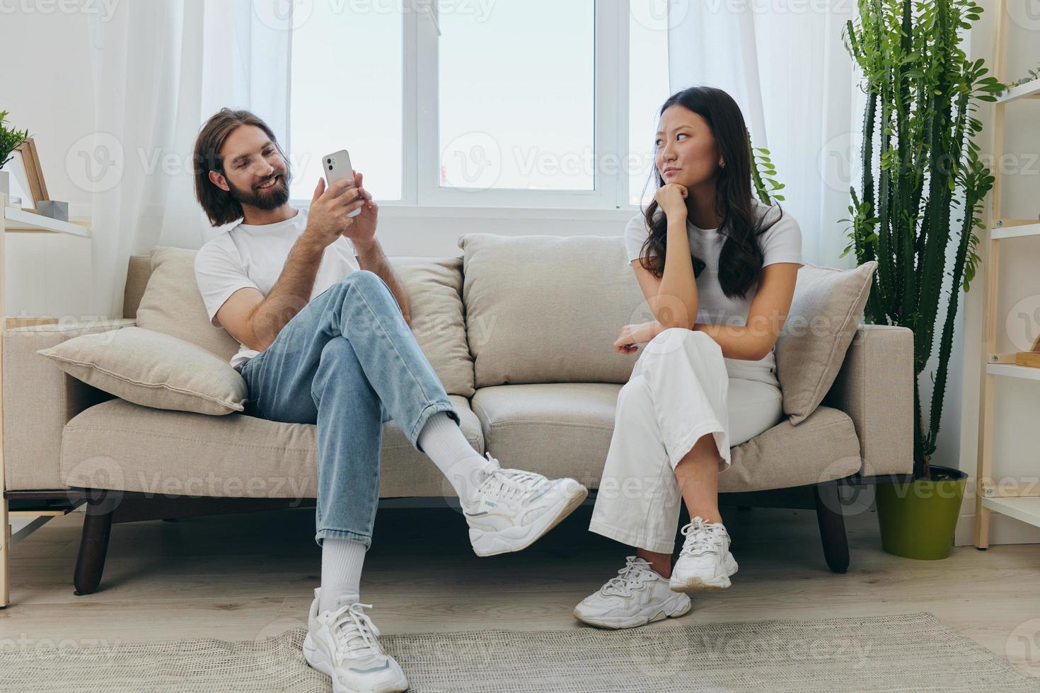A man looks at his phone screen and smiles during an argument with his girlfriend. The angry and hurt woman looks in his direction and is sad. Family discord at home, phone addiction photo