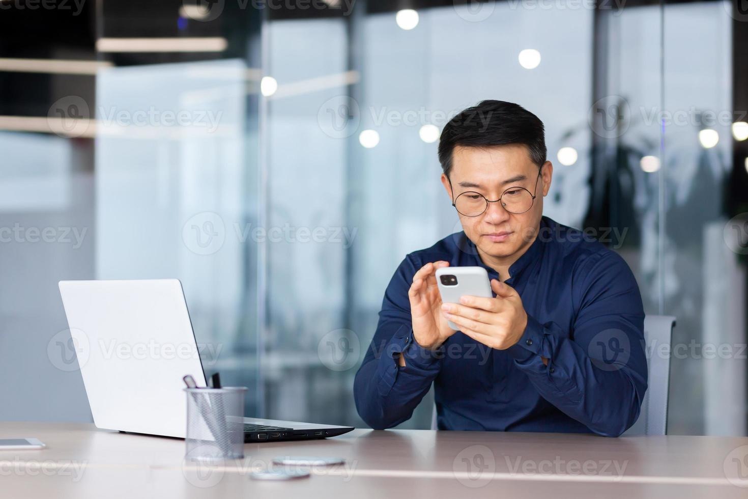 de cerca de grave y pensativo asiático empresario utilizando teléfono, hombre dentro oficina mecanografía mensaje, hojeada Internet paginas y acecho vídeo foto