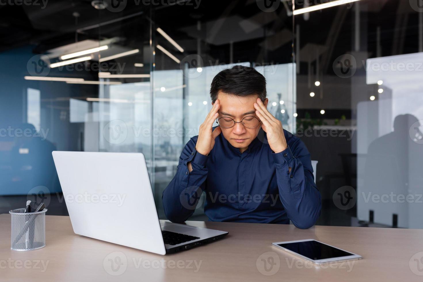 Stressed man working inside office, overworked asian man sitting at workplace sad and depressed holding hands on head, businessman frustrated thinking about problem photo