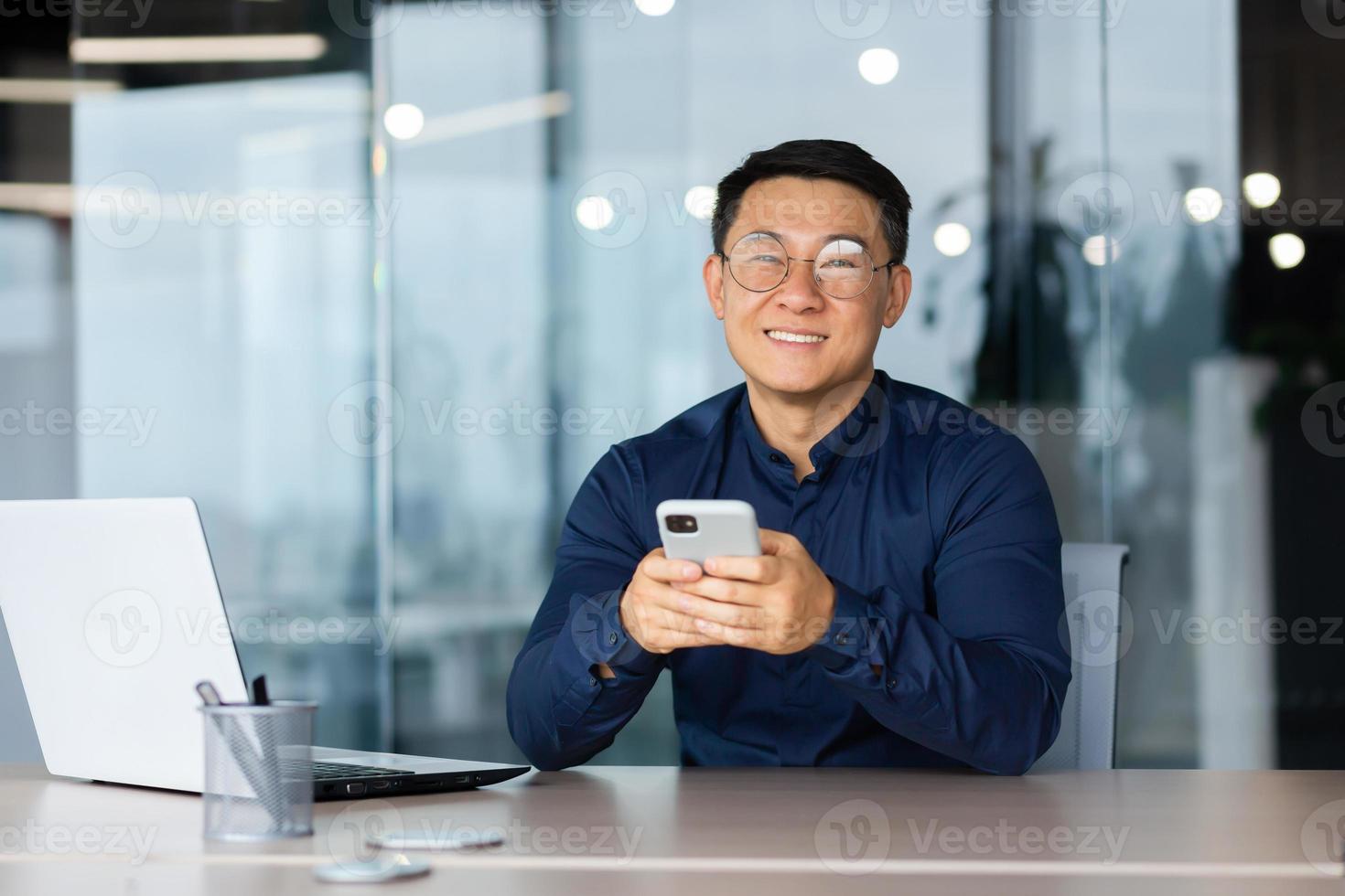 A young asian man sits in the office at a table, Holds a phone in his hands, smiles at the camera photo