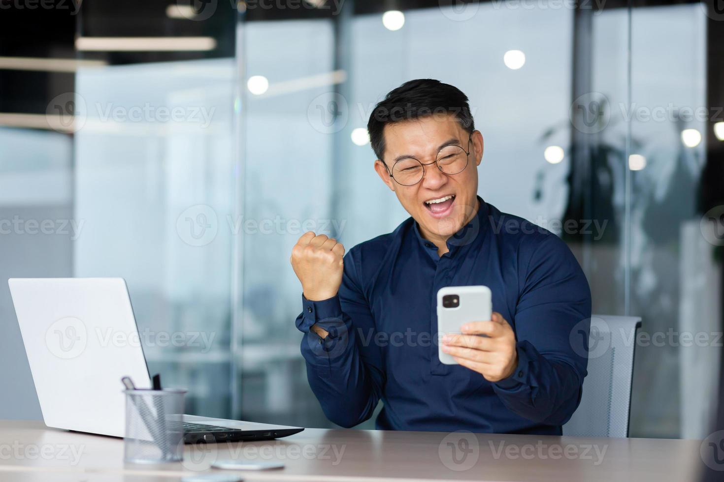 Successful asian businessman celebrating victory and happy good news notification from phone reading, man working inside office using laptop at work holding hand up triumph gesture photo