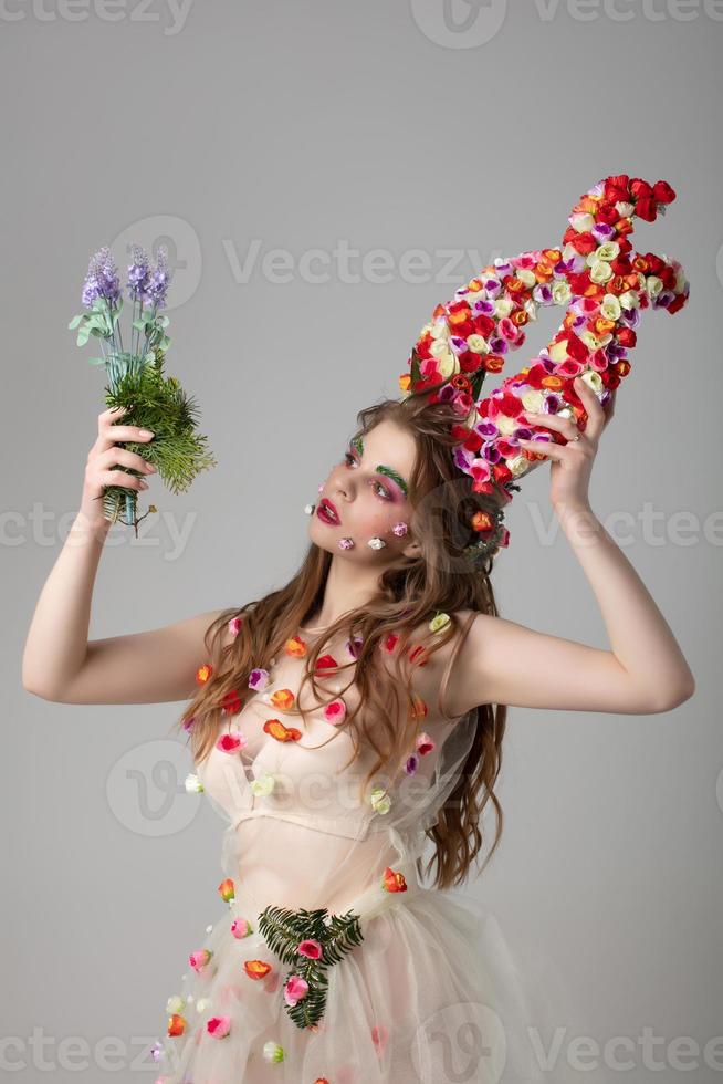 Fairy-tale girl model with floral horns and a forest bouquet on a studio background. Maleficent. Spring or summer beauty. Woman in the image of a flower. Goddess Flora photo