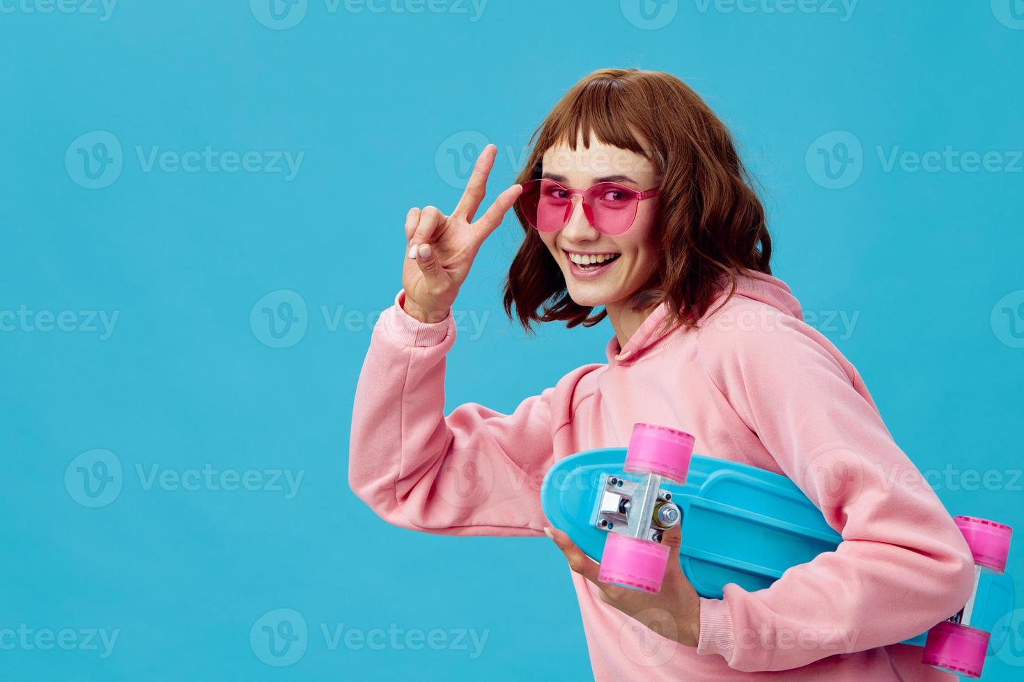 Funny joyful pretty redhead lady in pink hoodie sunglasses with penny board smiling posing isolated on blue studio background. Copy space Banner Offer. Fashion Cinema. Holiday activity photo