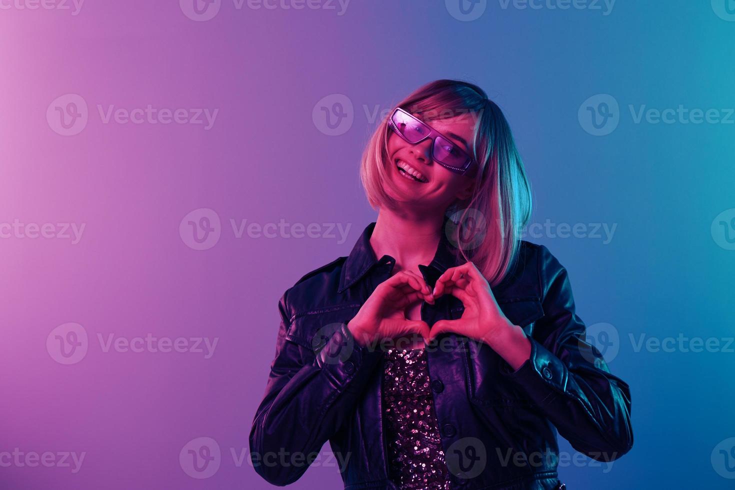 encantador hermosa rubia mujer en cuero chaqueta brillante vestir de moda Gafas de sol espectáculo corazón con dedos posando aislado en azul rosado color ligero estudio antecedentes. neón fiesta concepto. Copiar espacio foto